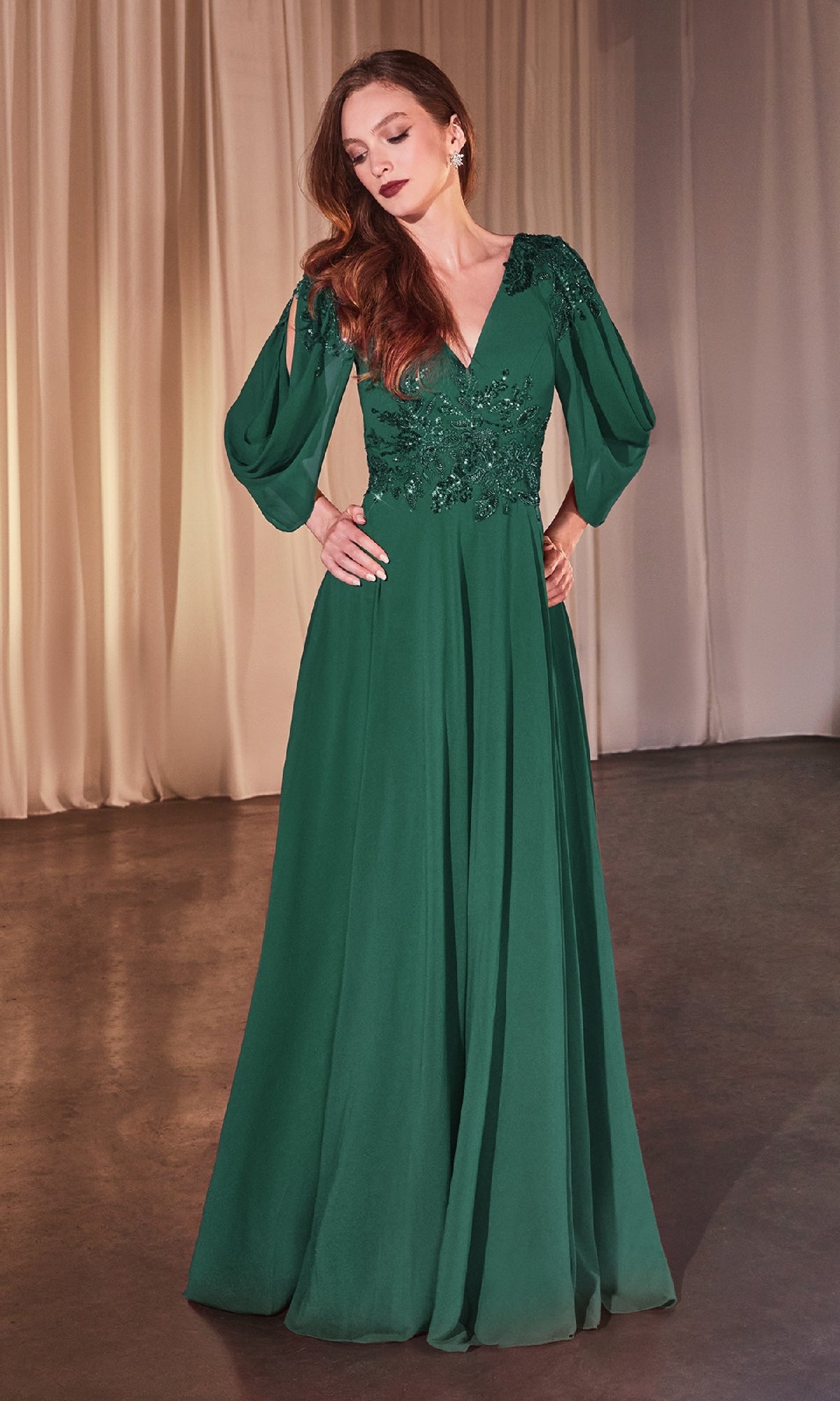 A woman stands indoors on a dark floor with cream curtains, wearing the Ladivine CDS508 Cowl-Sleeve Long Formal Dress in deep green, featuring sheer long sleeves and floral lace detailing on the bodice.